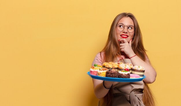 Giovane donna graziosa. fornaio di espressione pensando o dubitando con cupcakes