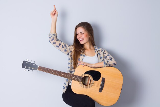 Giovane donna graziosa in camicetta beige e pantaloni neri che tiene una chitarra su sfondo grigio in studio