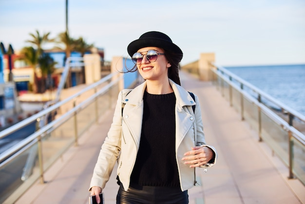 Giovane donna graziosa in cappello nero che cammina vicino al mare