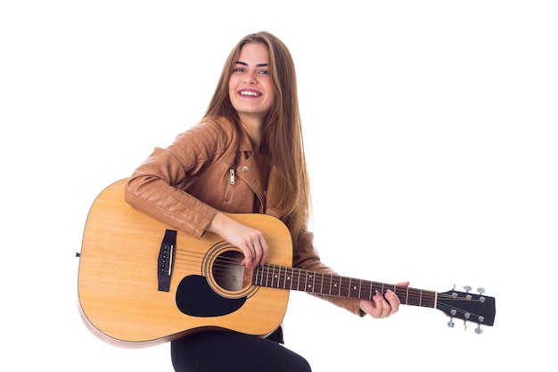 Giovane donna graziosa in giacca marrone e pantaloni neri in possesso di una chitarra su sfondo bianco in studio