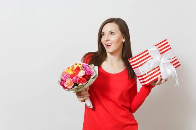 Giovane donna graziosa in vestiti rossi che tengono il mazzo di bei fiori delle rose, scatola attuale con il regalo isolato su fondo bianco. San Valentino, concetto di festa di compleanno della Giornata internazionale della donna