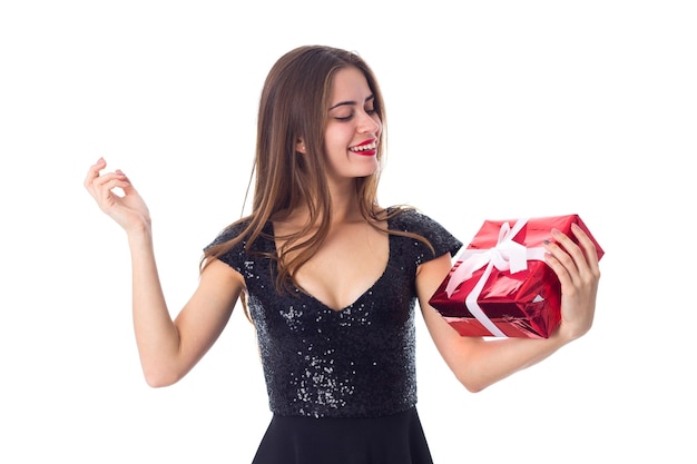 Giovane donna graziosa in vestito nero che tiene presente rosso e alza lo sguardo su sfondo bianco in studio