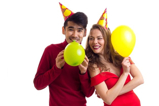 Giovane donna graziosa in vestito rosso e giovane uomo bello con cappelli colorati che soffia palloncini