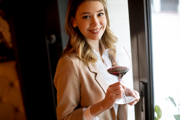 Giovane donna graziosa non-sposata con bicchiere di vino rosso che sta finestra vicina e che guarda da parte