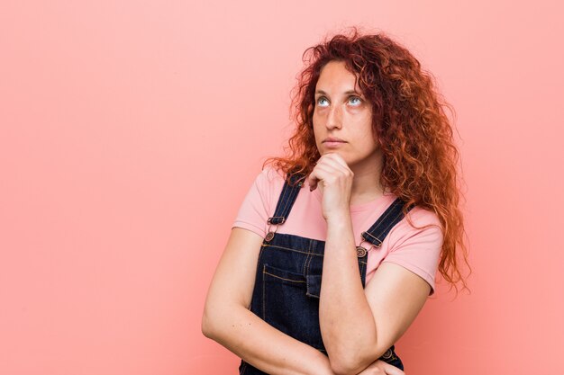 Giovane donna graziosa rossa dello zenzero che indossa un denim jeans guardando lateralmente con espressione dubbiosa e scettica.