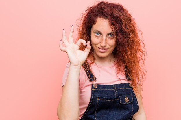 Giovane donna graziosa rossa dello zenzero che indossa un denim salopette allegro e fiducioso mostrando gesto ok.