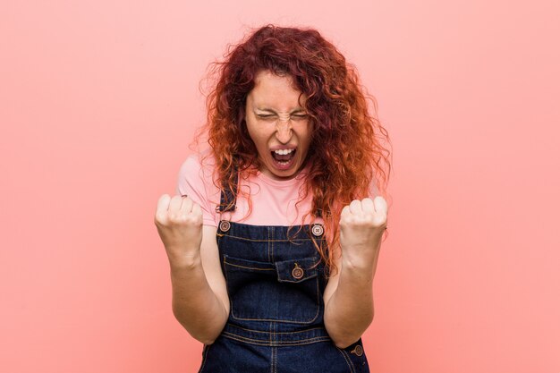 Giovane donna graziosa rossa dello zenzero che indossa una salopette di jeans tifo spensierato ed eccitato. Concetto di vittoria.