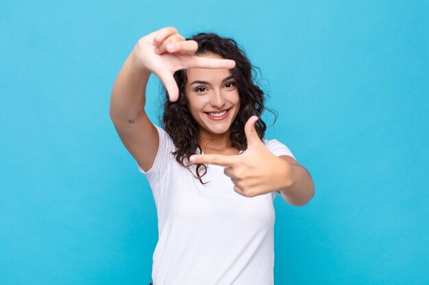 Giovane donna graziosa sentirsi felice, amichevole e positivo, sorridendo e facendo un ritratto o una cornice con le mani contro la parete blu