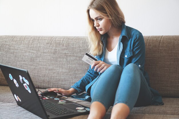 Giovane donna graziosa sicura che lavora con il computer portatile e la carta di credito a casa. Adesivi su un laptop senza etichette