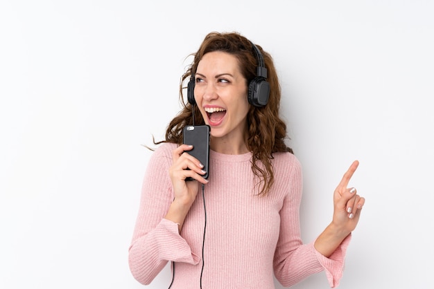 Giovane donna graziosa sopra isolata usando il cellulare con le cuffie e il canto