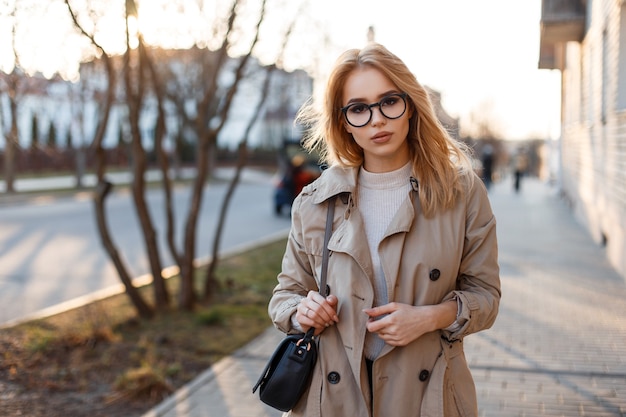 Giovane donna hipster in un elegante cappotto primaverile in occhiali alla moda in una maglietta bianca in una borsa di pelle nera che cammina lungo la strada su edifici d'epoca. Ragazza europea.