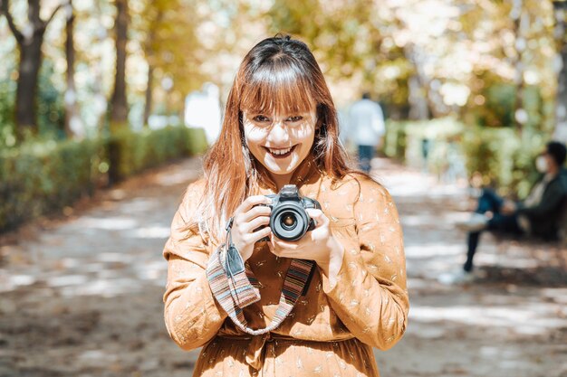 Giovane donna hipster su un vestito autunnale sorride alla macchina fotografica mentre scatta foto nel mezzo di un parco durante l'autunno