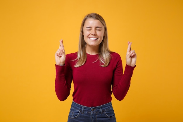 Giovane donna in abbigliamento casual in posa isolato su sfondo giallo arancione. Concetto di stile di vita delle persone. Simula lo spazio della copia. Aspettando un momento speciale, tenendo le dita incrociate, gli occhi chiusi, esprimendo un desiderio.