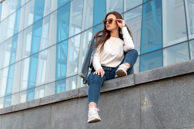 Giovane donna in abbigliamento giovanile alla moda e occhiali da sole Studentessa sta riposando sulla strada della città