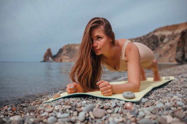 Giovane donna in abbigliamento sportivo beige con i capelli lunghi che pratica stretching all'aperto sul tappetino da yoga in riva al mare