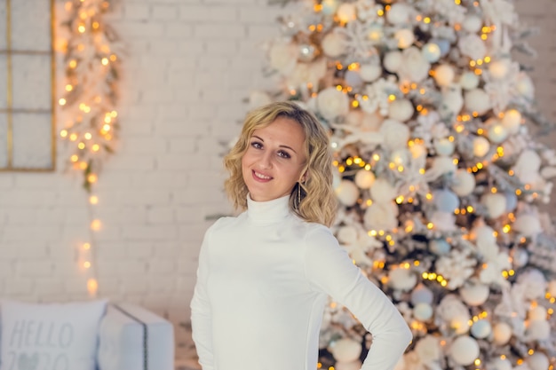 Giovane donna in abiti bianchi vicino all'albero di Natale con luci bokeh di fondo a Natale