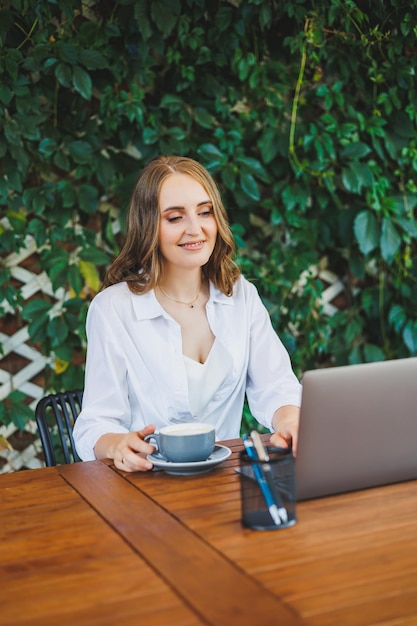 Giovane donna in abiti casual che beve caffè e parla al telefono lavorando su una terrazza verde davanti a un laptop guardando lo schermo e lavorando a un progetto a distanza dal lavoro in un bar