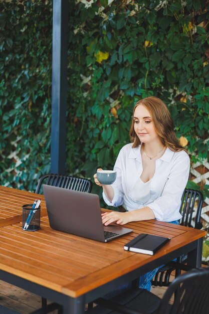 Giovane donna in abiti casual che beve caffè e parla al telefono lavorando su una terrazza verde davanti a un laptop guardando lo schermo e lavorando a un progetto a distanza dal lavoro in un bar