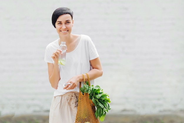 Giovane donna in abiti estivi leggeri con un sacchetto ecologico di verdure, verdure e una bottiglia d'acqua riutilizzabile. Stile di vita sostenibile. Concetto ecologico.