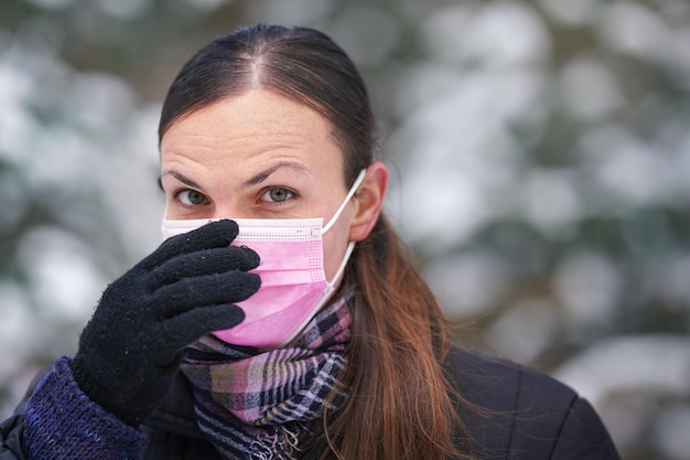 Giovane donna in abiti invernali caldi, indossando guanti di protezione, mettendo o rimuovendo la maschera monouso monouso per la bocca del virus del viso rosa, primo piano, sfocato sfondo di alberi innevati