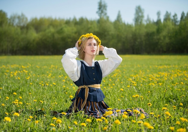 Giovane donna in abiti nazionali che indossa una corona di tarassaco gialla in campo primaverile Primavera