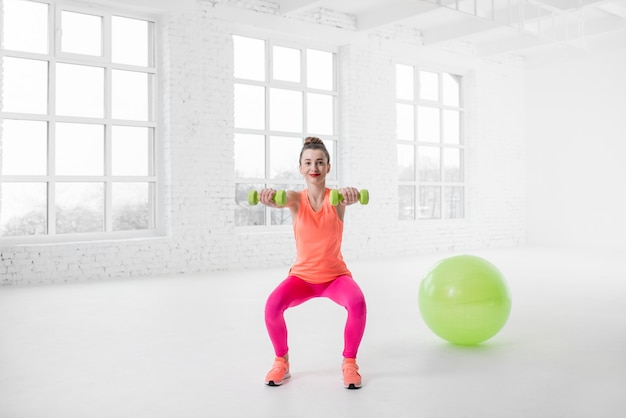 Giovane donna in abiti sportivi colorati accovacciata con manubri nella palestra bianca con palla fitness sul pavimento