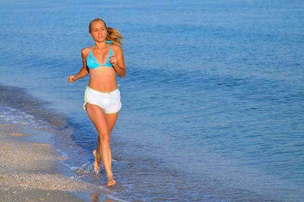 Giovane donna in abiti sportivi e scarpe da ginnastica in esecuzione vicino al bordo del mare e sorridente il giorno di estate pieno di sole. Felicità, vacanze e concetto di libertà