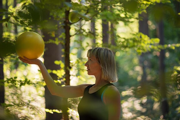 Giovane donna in abiti sportivi in piedi in una foresta illuminata dal sole che tiene palla pilates gialla
