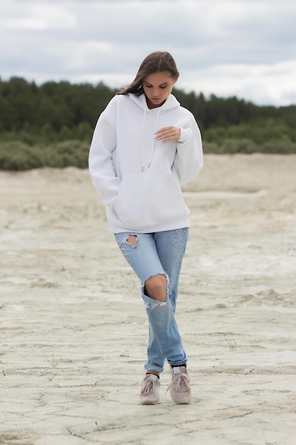 Giovane donna in abito alla moda che guarda la fotocamera e tocca la pancia sotto la felpa con cappuccio mentre si riposa sulla spiaggia in una giornata ventosa