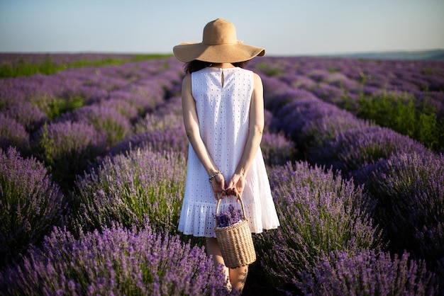 Giovane donna in abito bianco che tiene un cesto di paglia con fiori in piedi sul campo di lavanda