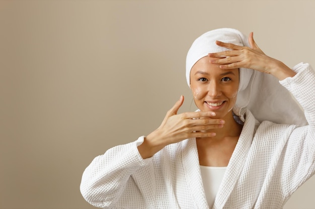 Giovane donna in abito bianco e con un asciugamano sulla testa versa l'applicazione di gel lozione o crema idratante per le mani dalla bottiglia bianca nel palmo della mano Cura del corpo sana dopo la doccia