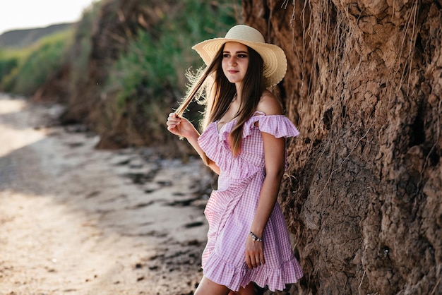 Giovane donna in abito e cappello di paglia cammina da sola sulla spiaggia di sabbia vuota al tramonto estivo sul mare e sorridente