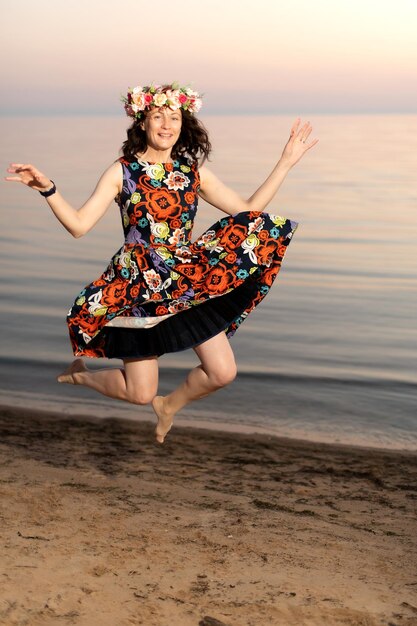 Giovane donna in abito fiorito con una corona di fiori in testa corre gioiosamente lungo la riva del mare