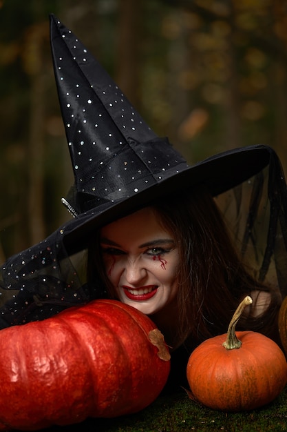 Giovane donna in abito nero con cappello da strega e zucca arancione disposta intorno nella foresta, concetto di Halloween