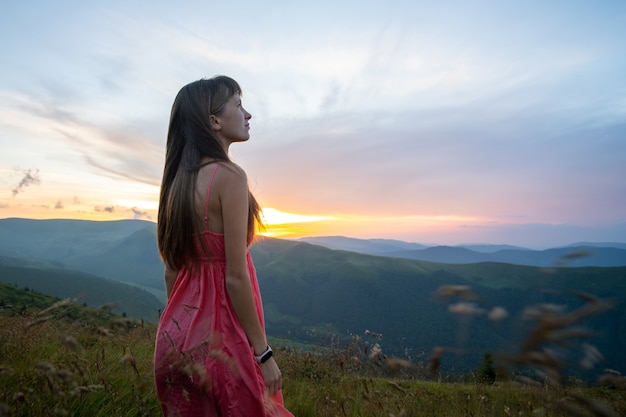 Giovane donna in abito rosso in piedi sul campo erboso in una serata ventosa in montagne autunnali godendo della vista della natura.