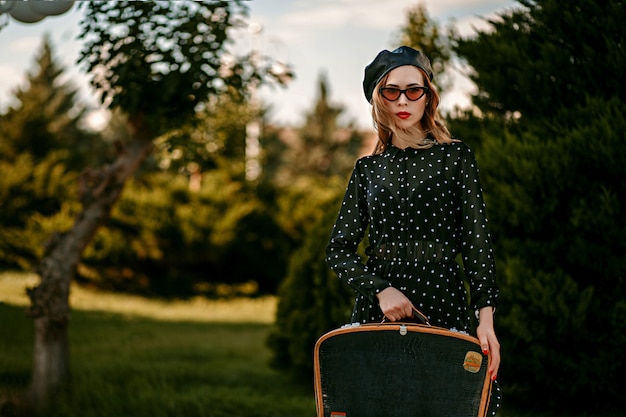 Giovane donna in abito vintage nero a pois con la retro valigia in mano in posa all'esterno