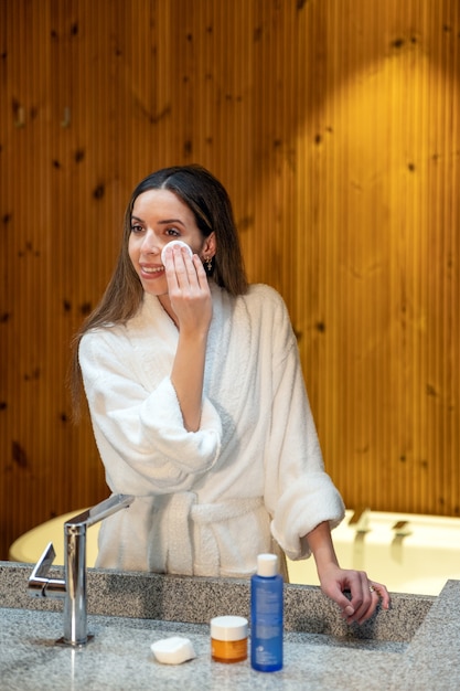Giovane donna in accappatoio bianco che pulisce la pelle del viso con un batuffolo di cotone e un prodotto cosmetico durante la procedura di bellezza quotidiana in bagno