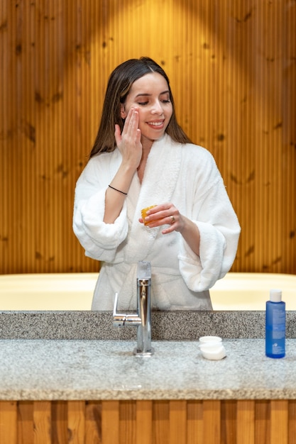 Giovane donna in accappatoio bianco in piedi davanti allo specchio in bagno e applicare la crema cosmetica sul viso durante la routine quotidiana di cura della pelle