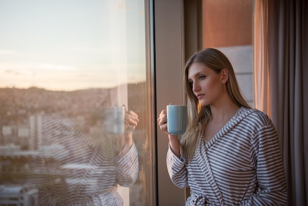 giovane donna in accappatoio che si gode il caffè serale e il bellissimo paesaggio al tramonto della città mentre si è in piedi vicino alla finestra