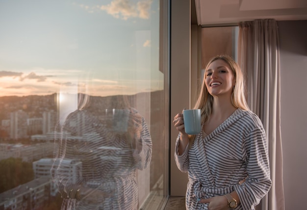 giovane donna in accappatoio che si gode il caffè serale e il bellissimo paesaggio al tramonto della città mentre si è in piedi vicino alla finestra
