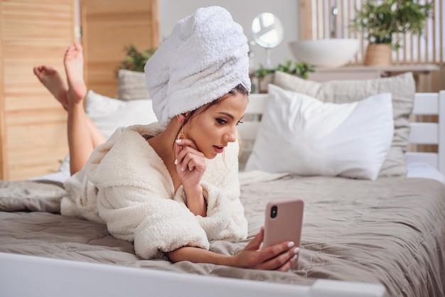 giovane donna in accappatoio e con un asciugamano sulla testa facendo selfie usando smartphone sul letto