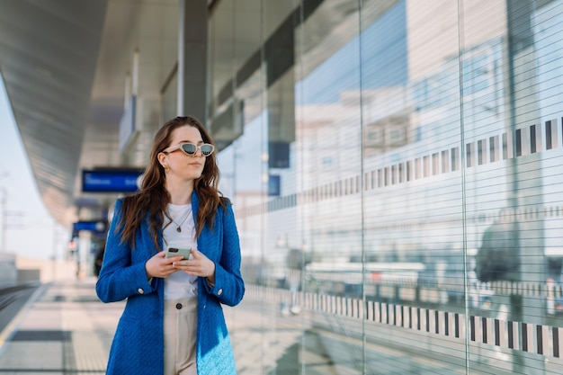 Giovane donna in attesa alla stazione ferroviaria Tenendo lo smartphone e aspettando il suo treno Trasporto pubblico