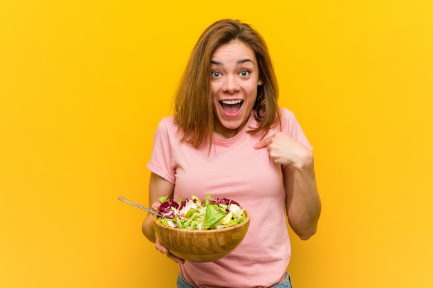 Giovane donna in buona salute che tiene un'insalata sorpresa indicando se stessa, sorridendo ampiamente