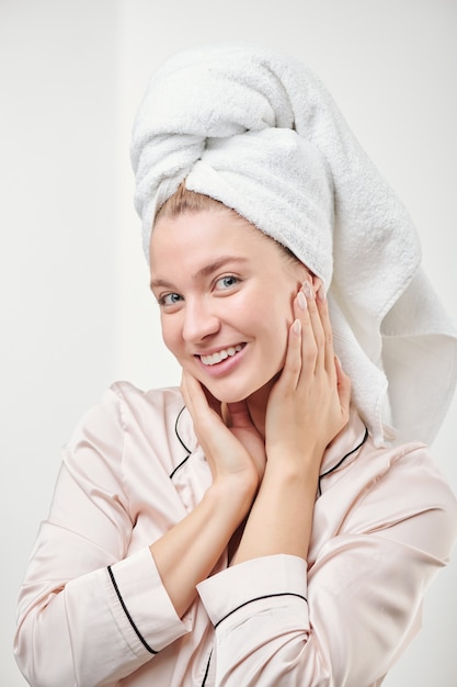 Giovane donna in buona salute con il sorriso a trentadue denti che tocca il suo viso luminoso e idratato mentre si gode la sua morbidezza