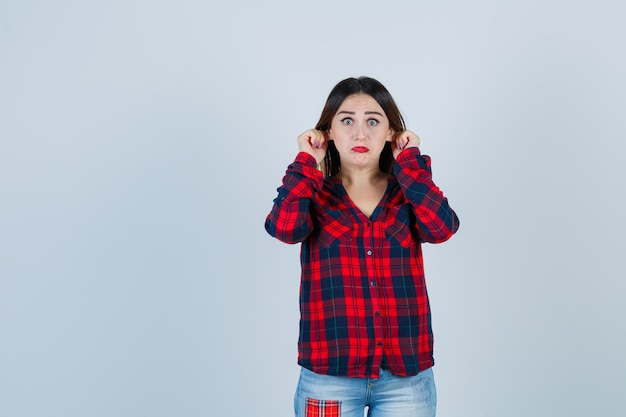 Giovane donna in camicia a quadri, jeans che tirano le orecchie e sembra spaventata, vista frontale.