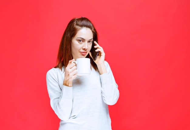 Giovane donna in camicia bianca che tiene una tazza da caffè bianca e uno smartphone nero e parla al telefono