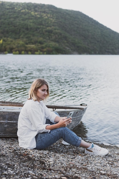 Giovane donna in camicia bianca e jeans con un telefono in mano si siede sulla barca sulla riva del lago di montagna
