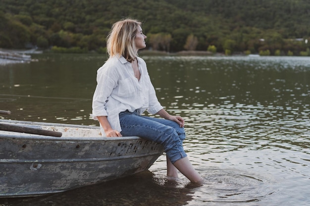 Giovane donna in camicia bianca e jeans si siede sulla barca e guarda il lago di montagna