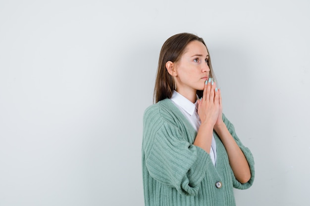 Giovane donna in camicia, cardigan con le mani in gesto di preghiera e sguardo fiducioso, vista frontale.