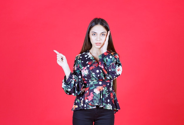 Giovane donna in camicia floreale in piedi sul muro rosso e mostra il lato sinistro